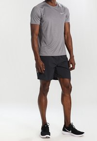 Graues Nike-T-Shirt mit kurzen Ärmeln, kombiniert mit schwarzen Sportshorts. Es hat einen runden Halsauschnitt, leichte Materialien und ein kleines Logo.