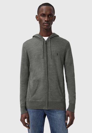 Mand iført grå hættetrøje med lynlås og forlommer samt blå jeans, stående foran en ensfarvet lysegrå baggrund.