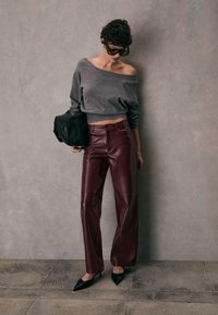 Grå off-shoulder sweater, højhalsede bordeaux læderbukser og sorte spidse hæle, med en sort taske og tekstureret stenvæg som baggrund.