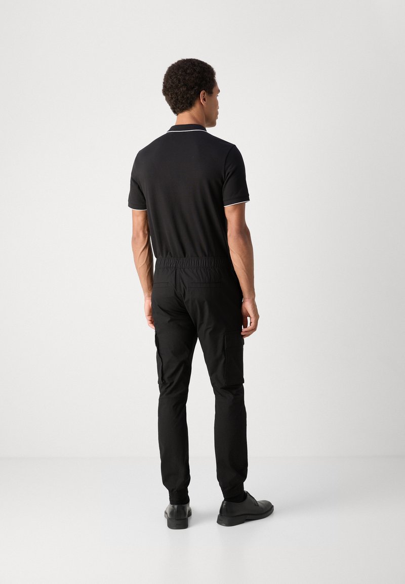 Polo shirt nero a maniche corte, con dettagli bianchi; abbinato a pantaloni cargo neri a gamba stretta e scarpe nere, visto da dietro.