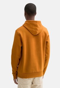 Felpa arancione con tasca a marsupio, orlo e polsini a coste, texture morbida e cappuccio sul retro. Indossata sopra pantaloni beige.