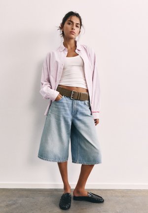 Jeune femme debout portant un débardeur blanc, une chemise rayée rose clair ouverte, un short en jean large avec une ceinture marron et des chaussures noires à enfiler.