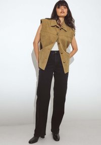 Kohvi nahast ilma varrukateta vest, millel on krae ja nupuga sulgus, koos kõrge vöökohtadega mustade pükste ja mustade pahkluu saapatega.