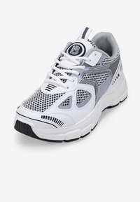 Zapatilla deportiva blanca con paneles de malla, detalles en gris y acentos en negro. Presenta una suela texturizada y un logo redondo en la lengüeta.