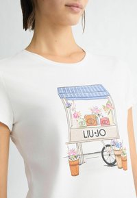 T-shirt en coton blanc présentant une illustration colorée d'un chariot à fleurs avec des sacs à main, le texte "LIU.JO" et des accents floraux dans des pots décoratifs.