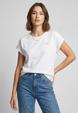 Junge Frau mit kurzen braunen Haaren, die ein weißes T-Shirt mit kleinem pinken Muschel-Logo und hoch taillierte blaue Jeans trägt, steht vor einem grauen Hintergrund.