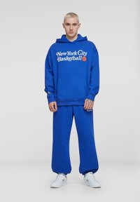 Niebieska bluza z napisem "New York City Basketball", z kieszenią typu kangur, w komplecie z dopasowanymi spodniami dresowymi. Oba elementy wykonane są z miękkiego materiału.