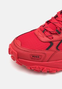 Chaussure de sport rouge avec tissu en maille texturée, semelle robuste, accents noirs et logo "HUGO" sur le côté près de l'orteil.