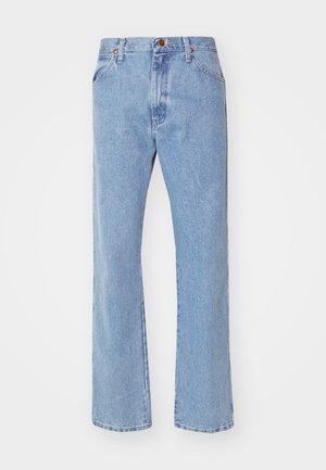Homme portant une chemise à carreaux bleue boutonnée, rentrée dans un jean bleu clair avec une ceinture beige et des bottes en daim marron, debout devant un fond blanc.