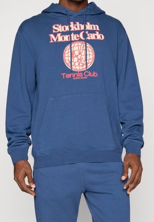 Mand iført en blå hættetrøje med pink tekst, der lyder "Stockholm Monte Carlo Tennis Club", og en globusgrafik foran.