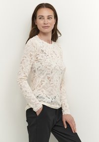 Top in pizzo a maniche lunghe di un crema chiaro, con motivi floreali, scollatura rotonda e orli delle maniche ondulati, abbinato a pantaloni scuri.