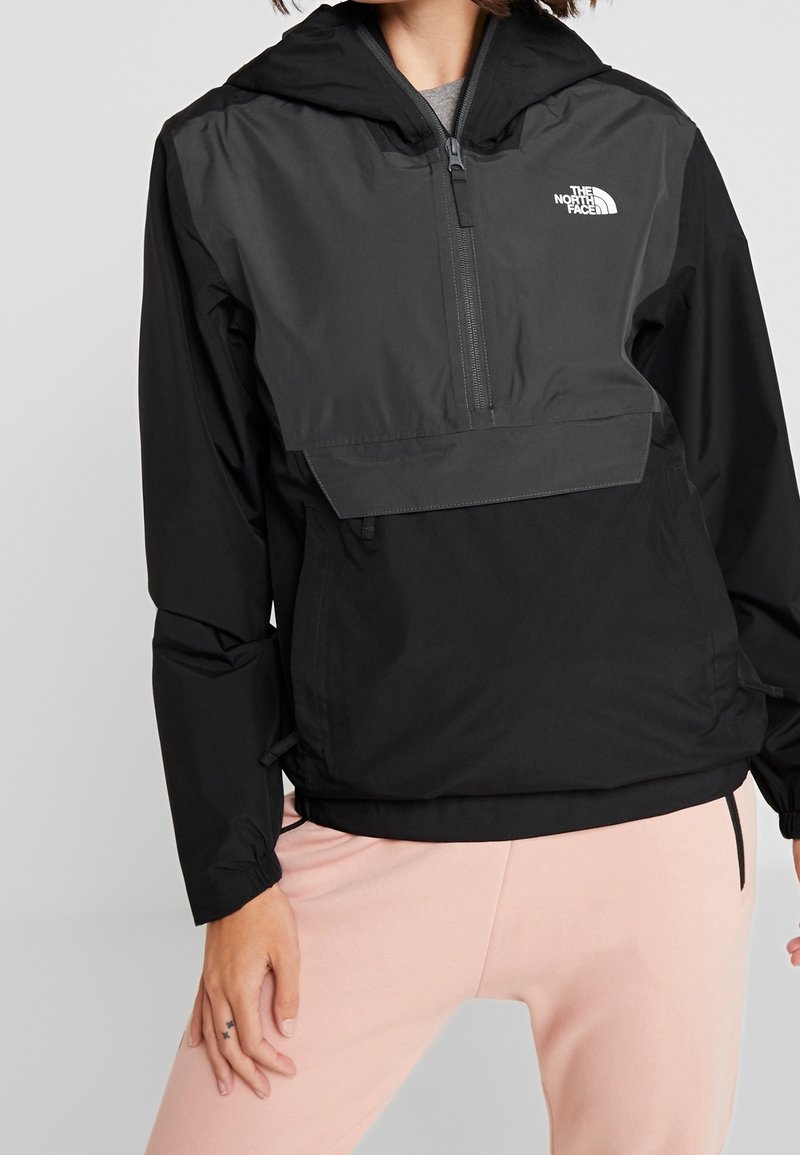 Personne portant une veste à capuche noire The North Face avec une fermeture éclair à l'avant et un pantalon rose clair sur un fond uni.