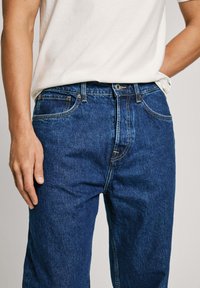 Pantalones vaqueros de mezclilla azul con un diseño de pierna recta, que presentan costuras doradas y un estilo clásico de cinco bolsillos. Llevados con una camiseta blanca.