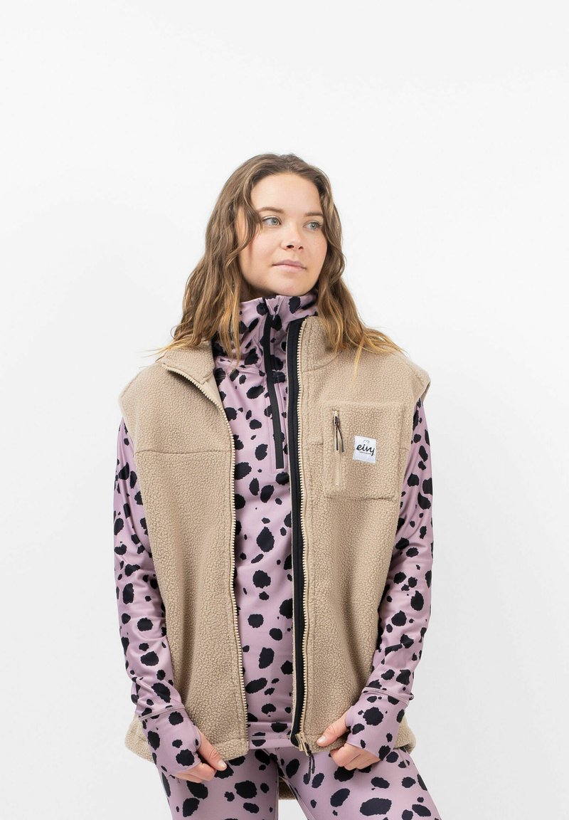 Beige Fleece-Weste mit Frontreißverschluss und Brusttasche, getragen über einem pinken Oberteil mit schwarzen Sprenkeln, ausgestattet mit langen Ärmeln mit Daumenlöchern.