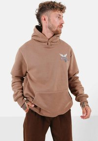 Brun fleece-huvtröja med en stor framficka, hög krage och broderad örn-design på bröstet. Mjuk textur, avslappnad passform.