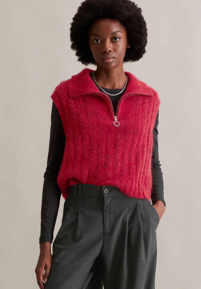 Rotes gestricktes ärmelloses Oberteil mit gerippter Textur und Reißverschlusskragen. Getragen über einem schwarzen Langarmshirt und kombiniert mit dunkelgrauen Hosen.