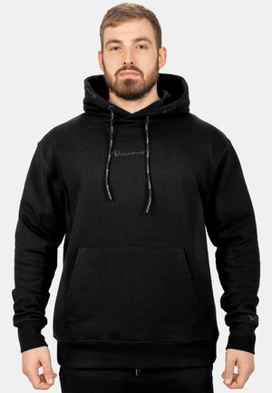 Mann mit kurzen Haaren und Bart, der einen schwarzen Hoodie mit Fronttasche und Kordelzug trägt, vor einem einfarbigen hellen Hintergrund stehend.