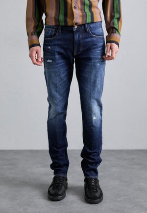 Tumed sinised teksad slim-fit lõikes, millel on kulumise üksikasjad ja õrn tuhmumine. Sobitatud mustade tossudega ja mitmevärvilise triibulise särgiga.