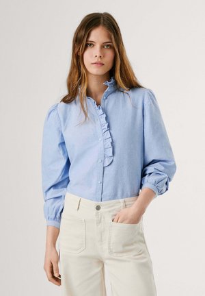 Blusa di cotone azzurro chiaro con parte anteriore arricciata, chiusura a bottoni e maniche a sbuffo. Abbinata a pantaloni bianchi con tasca.