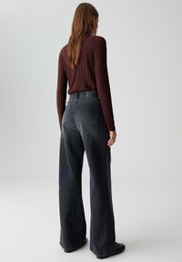 Femme aux longs cheveux bruns portant un haut à manches longues côtelé rouge foncé et un jean large taille haute noir délavé, se tenant de dos.