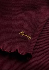 Burgunderfarbener, gerippter Stoff mit gewelltem Rand und goldbesticktem "Superdry"-Logo an der Seite. Weiche Textur, Nahaufnahme.
