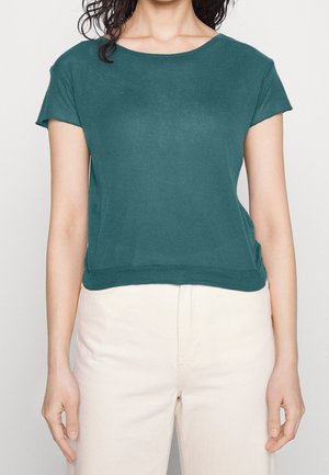 Turkusowa koszulka z krótkim rękawem i okrągłym dekoltem, o luźnym kroju, wykonana z miękkiego materiału, w połączeniu z jasnymi spodniami.