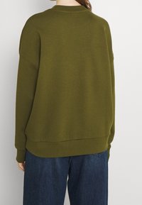 Olivekowo zielona, oversize bluza z okrągłym dekoltem i opadającymi ramionami. Miękka tkanina z prążkowanym wykończeniem. W zestawie z ciemnoniebieskimi spodniami z szerokimi nogawkami.