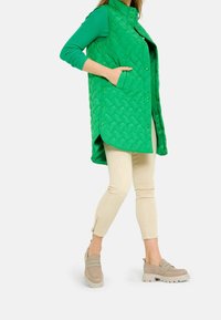 Chaleco acolchado verde con cuello alto, botones a presión y bolsillos laterales, combinado con pantalones de color beige claro y zapatos tipo slip-on color topo.