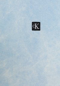 Světle modrá bavlněná látka s jemnou texturou; uprostřed je černé čtvercové logo s bílým nápisem "cK".