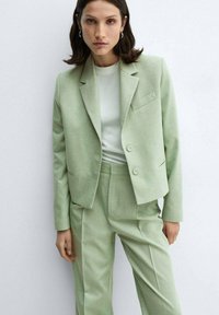Light green tailored suit with a collared jacket and matching trousers. Soft fabric, minimalist design, single chest pocket, two buttons.