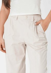 Lichtbeige broek met een elastische tailleband, zijzakken, knoopdetail en een subtiel logo op de dij. Glad materiaal met een relaxte pasvorm.