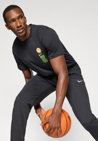 Sort bomulds T-shirt med basketball grafik og grønne design; kombineret med sorte bukser, hvor Nike-logoet vises. Holder en orange basketball.