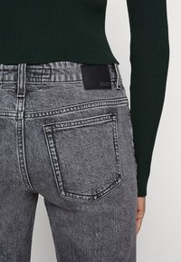 Jeans de denim negro con textura desgastada, cuentan con bolsillos traseros y un parche de cuero negro en la cintura, combinados con una camiseta de canalé verde oscuro.