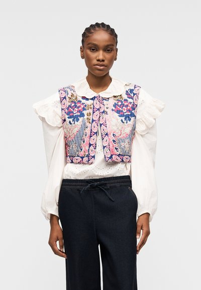Femme portant un chemisier blanc à manches volantées et un gilet brodé de motifs floraux, associé à un pantalon sombre à cordon, debout devant un fond neutre.