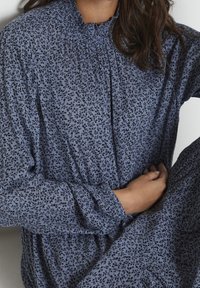 Mujer vistiendo un vestido largo de manga larga con un patrón floral azul, sentada con las manos suavemente entrelazadas en el regazo, contra un fondo liso.