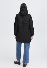 Doudoune noire avec un motif en losange, dotée d'une capuche et de manches longues, assortie à un jean bleu ample et des chaussures sombres.
