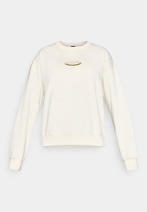 Hanorac crem cu guler rotund, umeri căzuți și manșete riburate. Prezintă o etichetă cu logo auriu în centru. Textură moale a țesăturii.