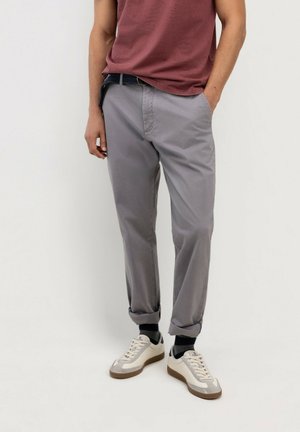 Hombre con pantalones grises remangados, calcetines oscuros, zapatillas blancas y una camisa granate, con una mano en el bolsillo, sobre un fondo liso.