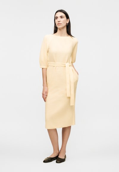 Γυναίκα με μακριά σκουρόχρωμα μαλλιά, που φοράει ένα ανοιχτό κίτρινο φόρεμα μήκους μέχρι το γόνατο, με μανίκια τριών τετάρτων, ζώνη στη μέση και μαύρα μπαλαρίνα παπούτσια, στέκεται μπροστά σε λευκό φόντο.