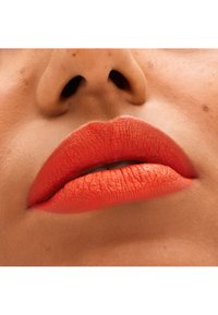 Rouge à lèvres orange mat appliqué sur des lèvres pleines avec une texture fine et légèrement craquelée, sur un fond de peau lisse aux tons chauds.