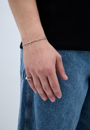 Ezüst lánc karkötő kereszt medállal. A kéz farmer anyagon helyezkedik el, mutatva egy gyűrűt az ujján. Texturált és minimalista dizájn.