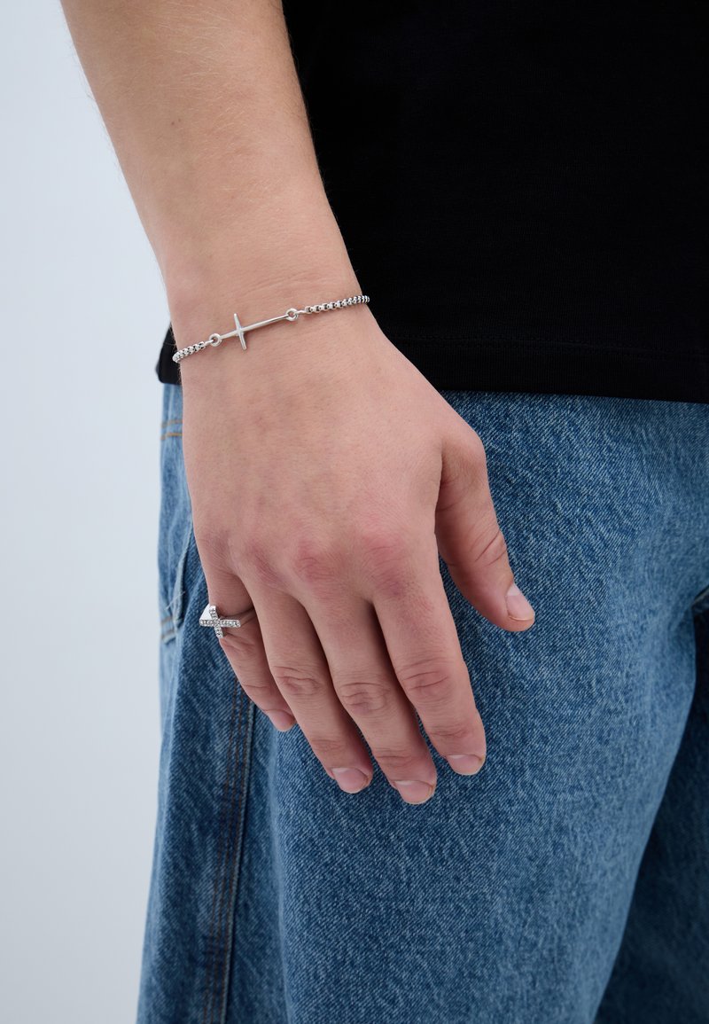 Brățară din lanț argintiu cu pandantiv în formă de cruce. Mâna este așezată pe un material denim, arătând un inel pe deget. Design texturat și minimalist.