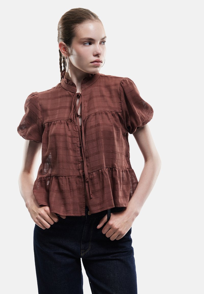 Blusa de color óxido con tejido texturizado, mangas cortas abullonadas, cierre frontal con botones y dobladillo tipo peplum. La blusa se ata en el escote.