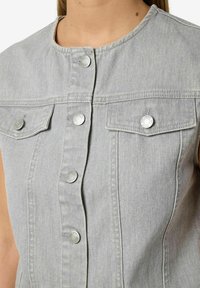 Siva traper bez rukava s okruglim izrezom, srebrnim dugmadi na kopčanje i dva džepa na prsima s glatkom, teksturiranom površinom.