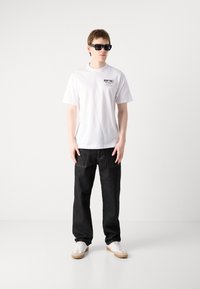 Hvit kortermet t-skjorte med logo, i kombinasjon med løse svarte jeans og hvite sneakers med lysebeige såler og detaljer.