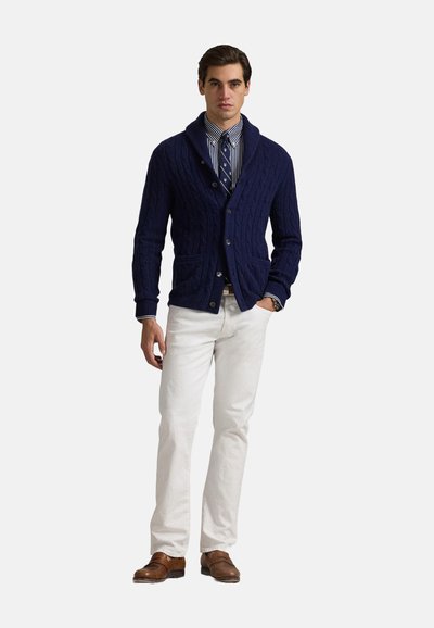 Cardigan in maglia a trecce blu navy con colletto sciallato, abbottonato, indossato sopra una camicia a righe e una cravatta, abbinato a pantaloni bianchi e scarpe eleganti marroni.