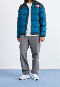 Person i en blå og sort pufferjakke, hvid skjorte, løse grå jeans og hvide sneakers, der står på en blå gulv mod en hvid væg.