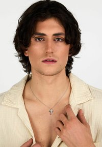 Collana a croce argentata con accenti trasparenti, indossata su un petto nudo, sopra una camicia beige testurizzata con colletto. Capelli naturali e mossi.