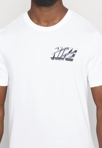 Camiseta de algodón blanca con un diseño del logotipo "NIKE" en plateado metálico en el lado izquierdo del pecho, cuello redondo y mangas cortas.