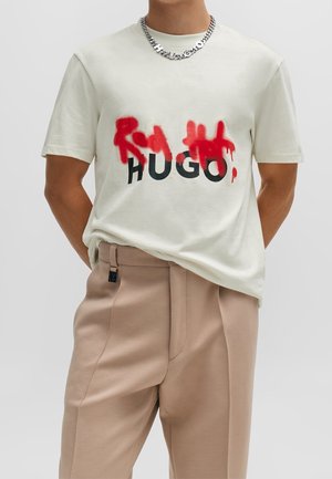 T-shirt à manches courtes crème avec texte rouge de style graffiti sur "HUGO" en gras noir, associé à un pantalon beige clair.